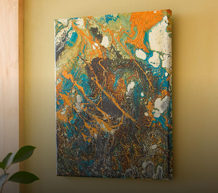 An abstract canvas wall art print featuring a fluid marble pattern in vibrant orange, teal blue, white, and dark gray, hanging on a warm beige wall.