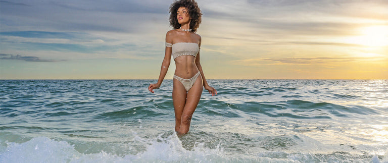 A woman stands in shallow ocean water at sunset wearing a light beige off-the-shoulder bikini. The background shows blue-green waves, a cloudy sky, and warm golden light near the horizon.