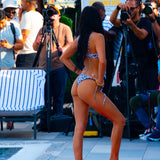 A woman posing from behind in a brown ribbed bikini on a red carpet, with photographers and a lounge chair in the background.